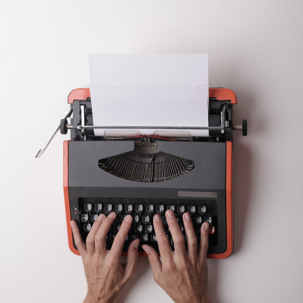 Two hands on the keys of an orange portable manual typewriter with a blank sheet of paper.